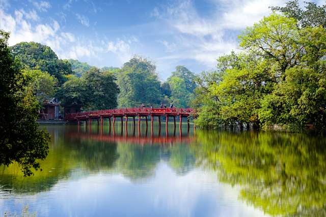 People on the red Huc Bridge entrance to Ngoc Son temple on Hoan Kiem lake by green trees, Hanoi, Vietnam