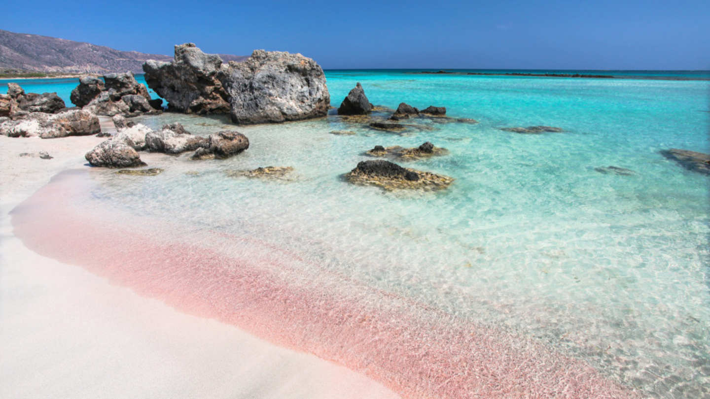 A view of the pink sand in shallow blue water by large rocks at Elafonisi Beach, Crete, Greece