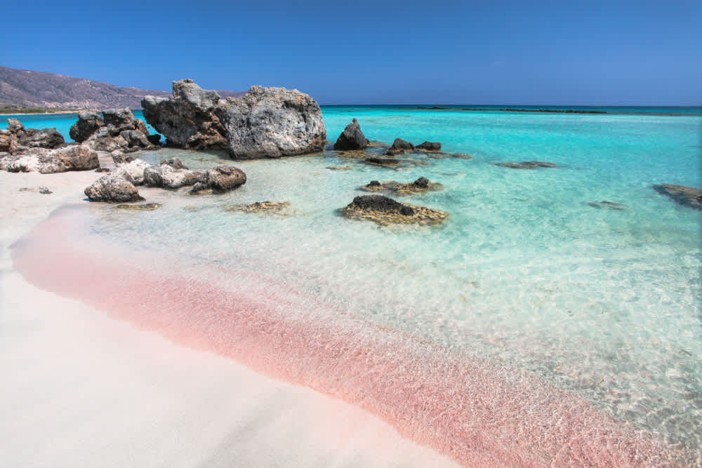 A view of the pink sand in shallow blue water by large rocks at Elafonisi Beach, Crete, Greece