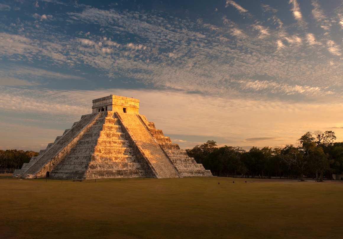 A view of the El Castillo pyramid in front of green trees at sunset, Chichén Itzá, Mexico