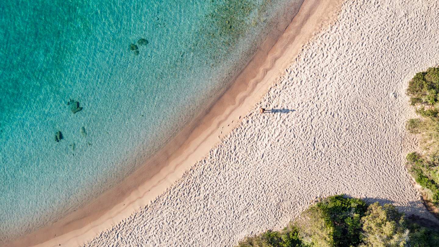 A bird's eye view of a person standing alone by the clear sea on a large sandy beach, Sardinia, Italy