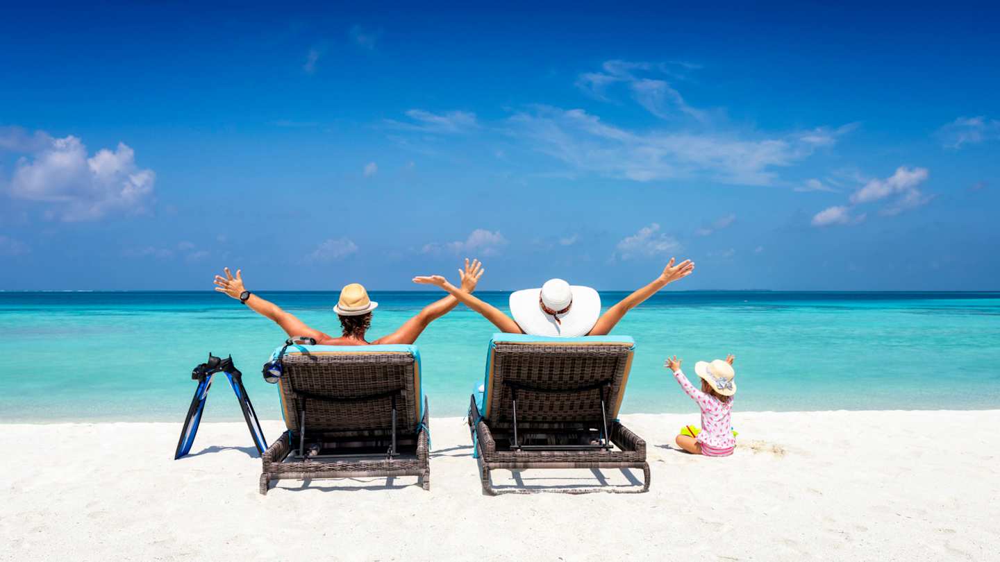 A young family sitting on sun loungers looking out to sea on a sandy beach in the sun