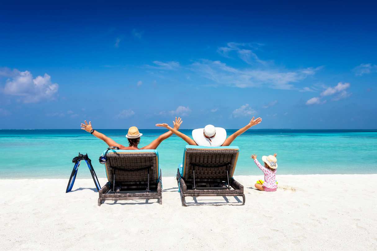 A young family sitting on sun loungers looking out to sea on a sandy beach in the sun