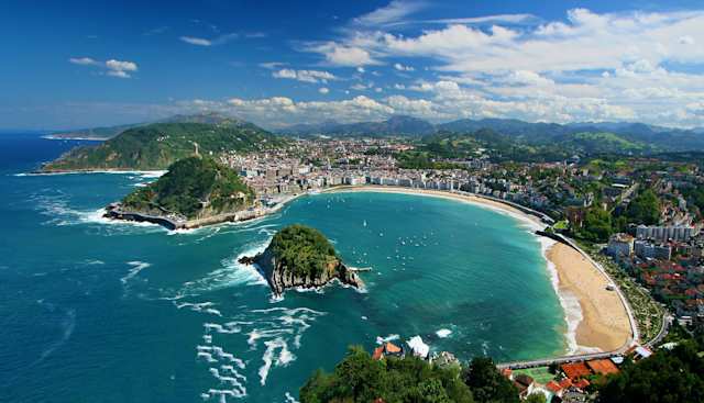 Ondaretta Beach alongside La Concha Beach, with waves rolling up the sand from the blue sea on a sunny day and buildings and mountains in the background, San Sebastian, Spain
