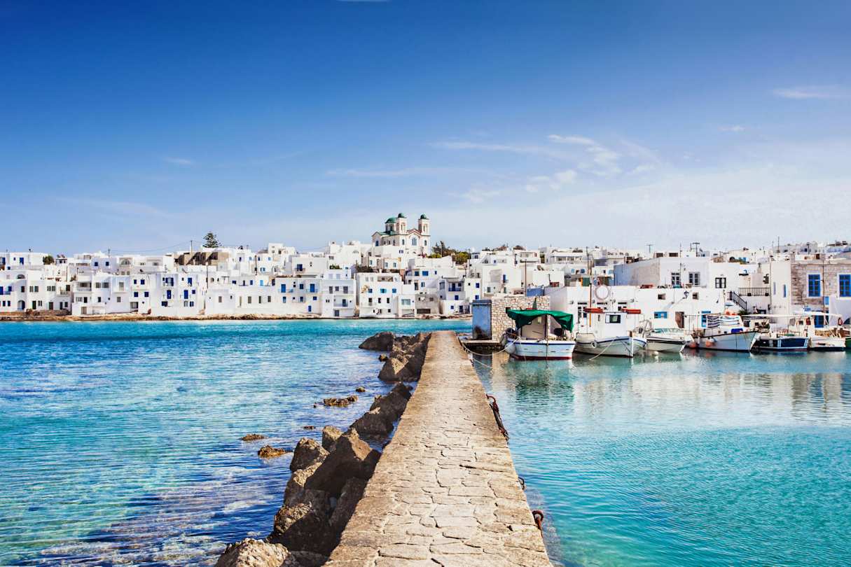 A brick pier jutting out into the blue sea next to docked boats at the harbour in Naoussa, Paros, Greece