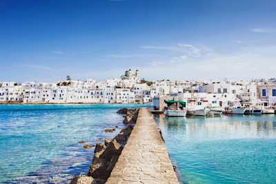 A brick pier jutting out into the blue sea next to docked boats at the harbour in Naoussa, Paros, Greece