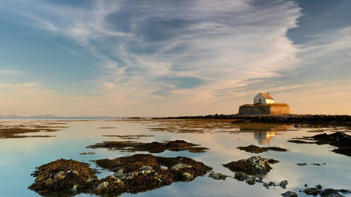 A view of St Cwyfan’s Church on a hill in the distance behind water and rocks at sunset in Anglesey, Wales, UK
