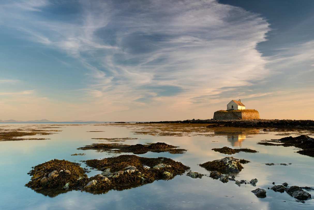 A view of St Cwyfan’s Church on a hill in the distance behind water and rocks at sunset in Anglesey, Wales, UK