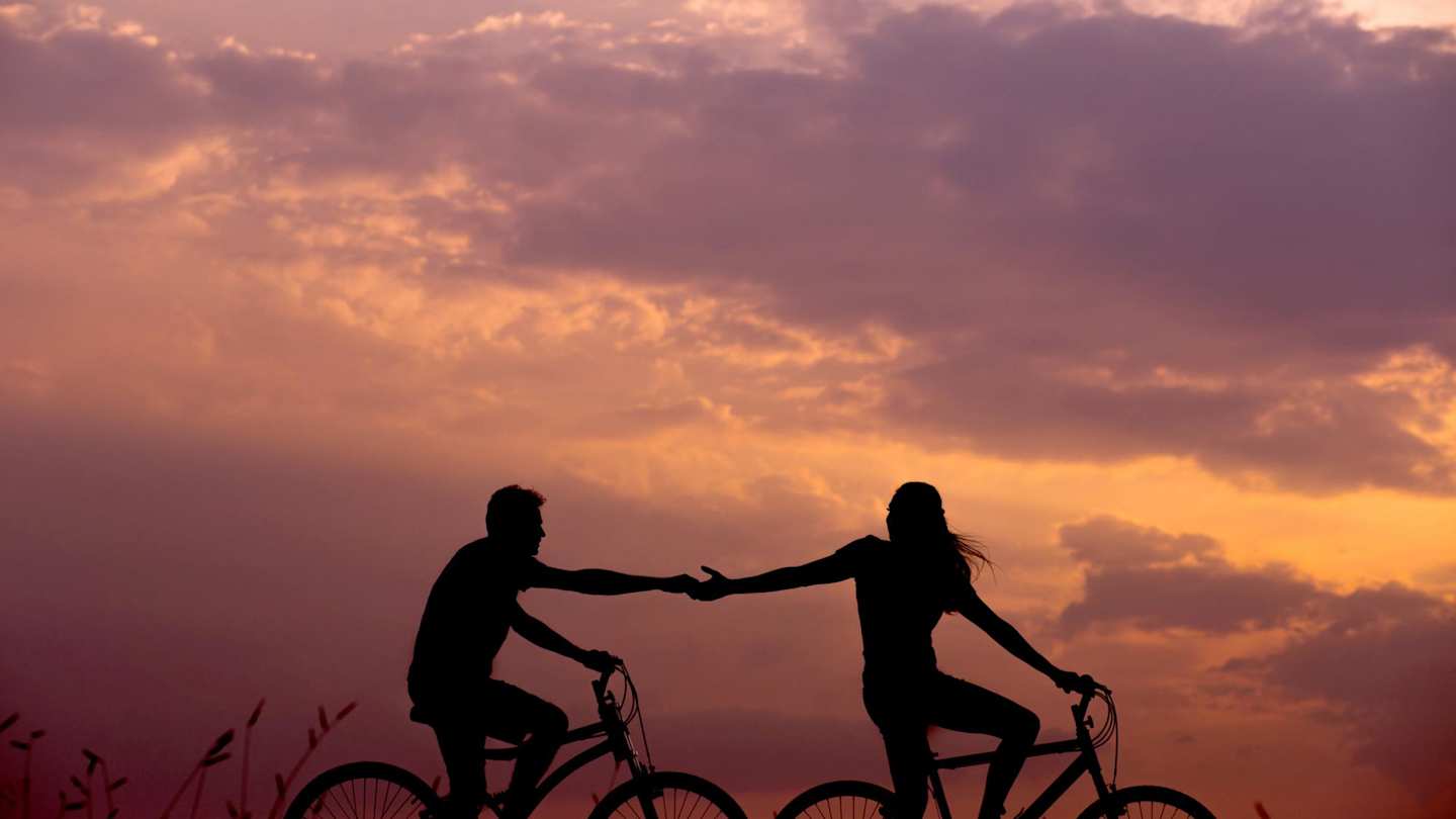 Two silhouetted people holdings hands while on bicycles at sunset