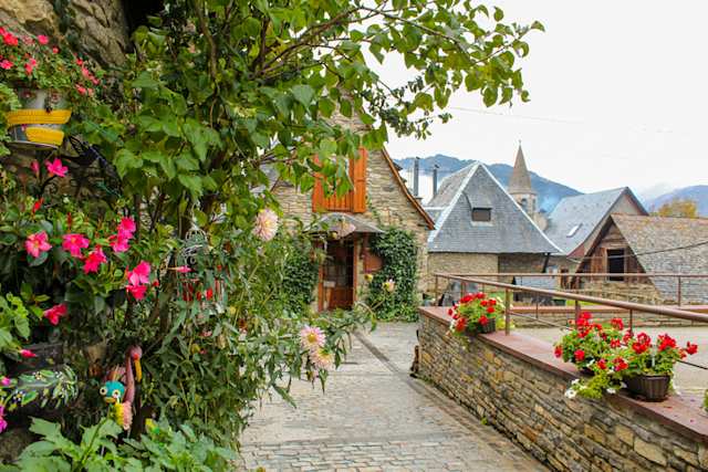 Pink flowers and vines along cobbled pathway with stone houses in distance, Bagergue, near Baqueira-Beret