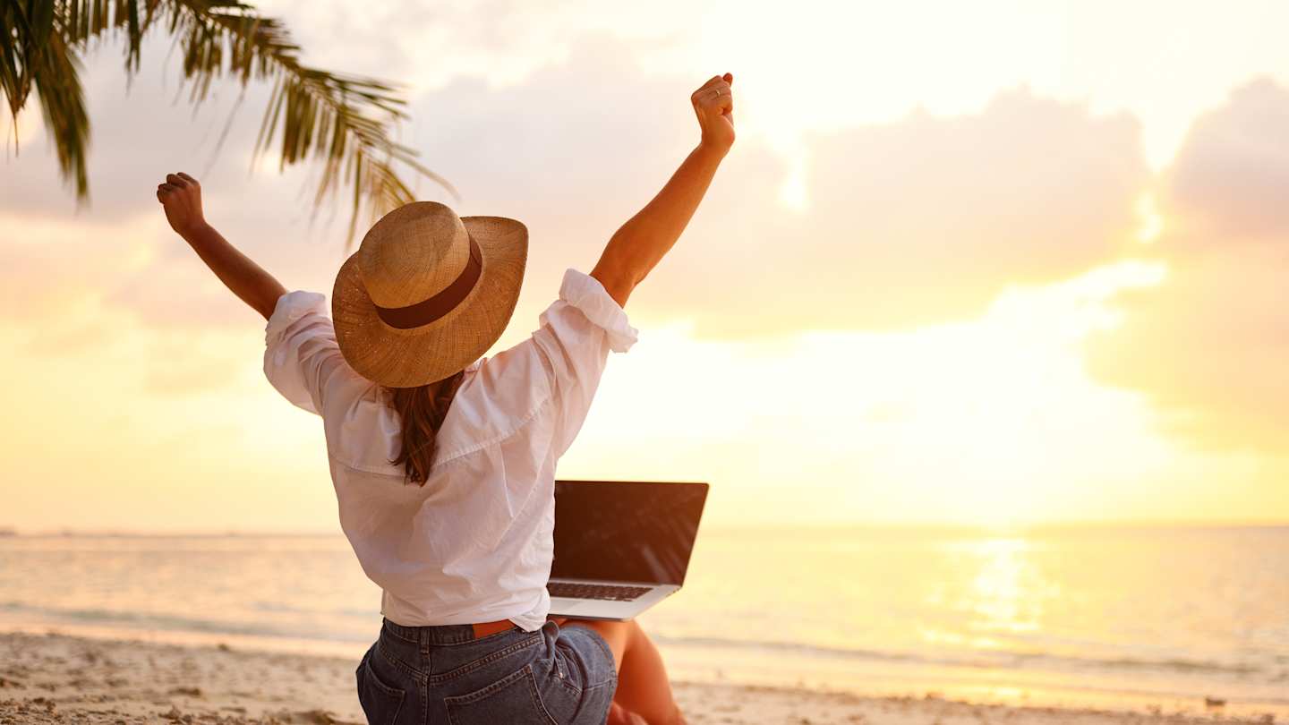 A woman celebrating with her laptop while working remotely on a beach as a digital nomad