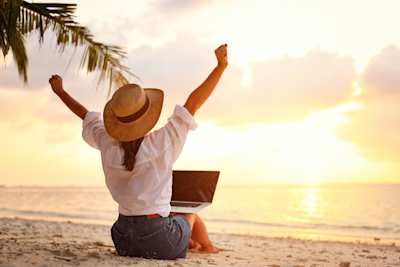 A woman celebrating with her laptop while working remotely on a beach as a digital nomad