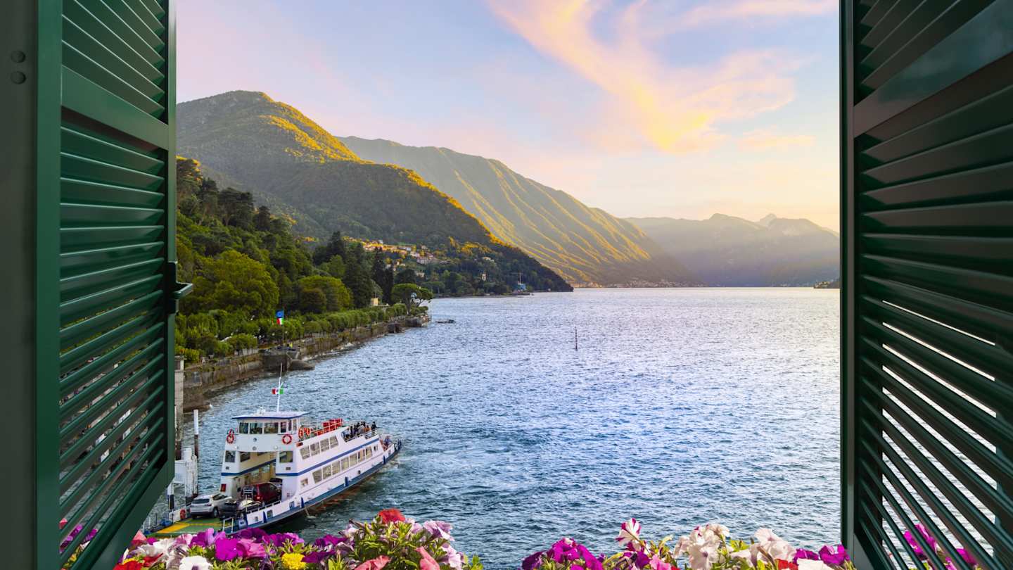 View of mountains and ferry boat at sunset from a terrace with flowers and shutters in Bellagio, Lake Como, Italy