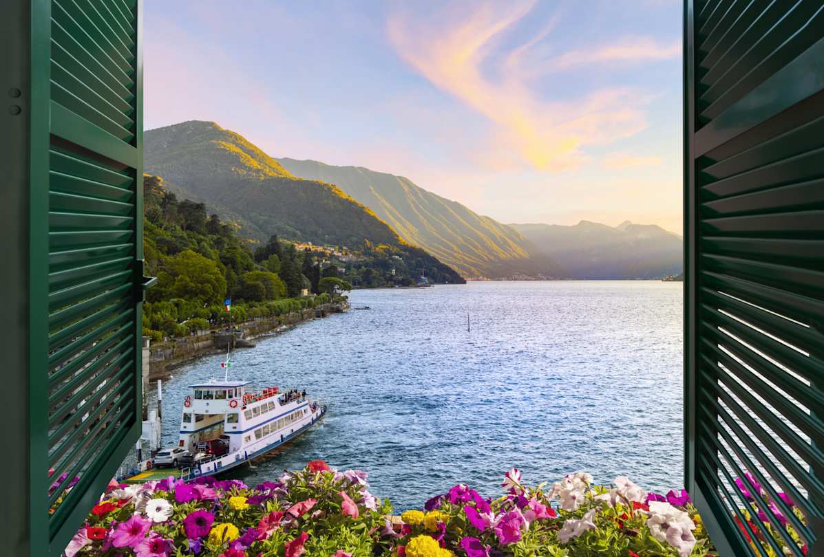 View of mountains and ferry boat at sunset from a terrace with flowers and shutters in Bellagio, Lake Como, Italy