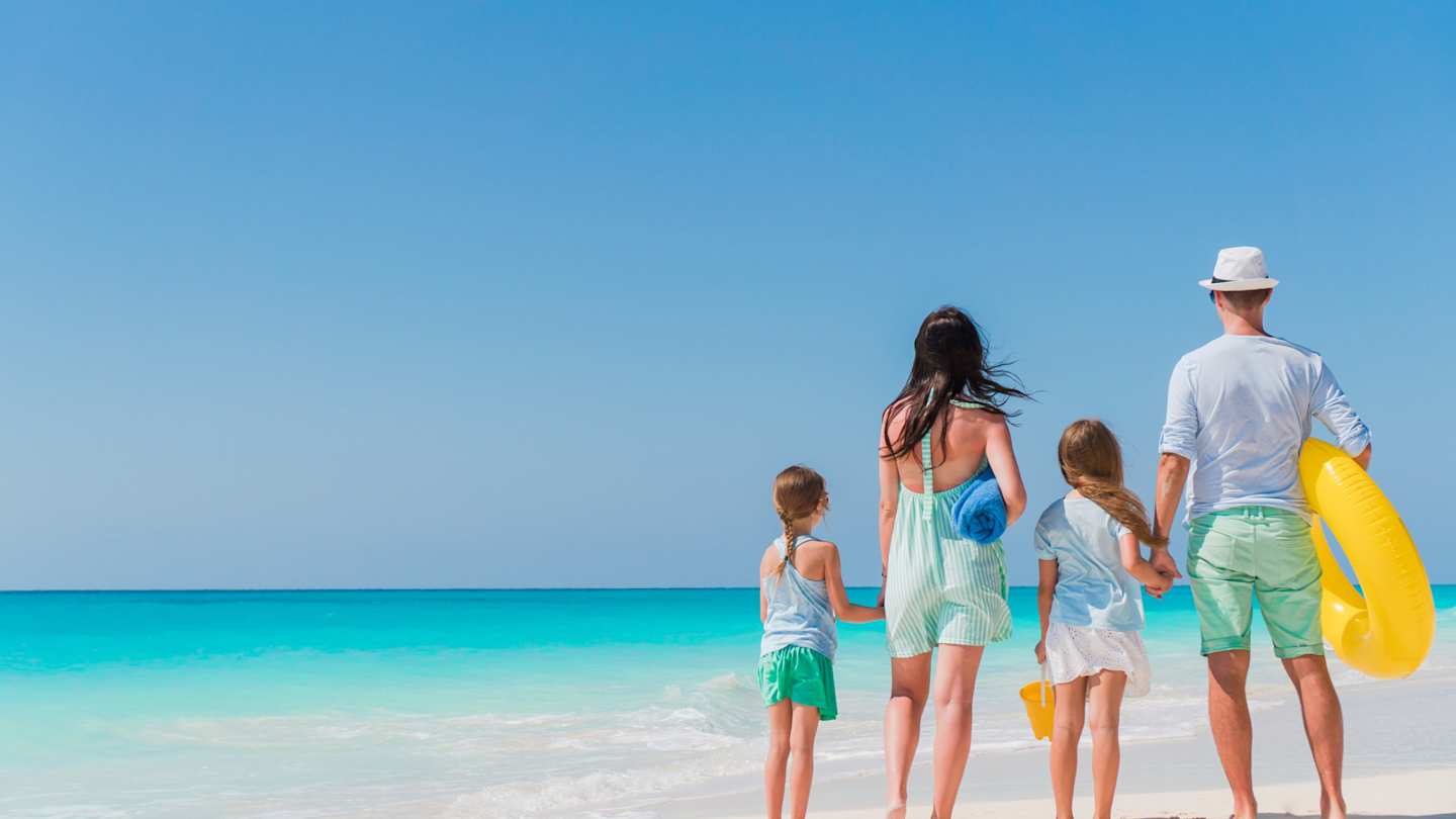 A young family standing on a sandy beach looking out to the clear blue sea on a hot day in summer
