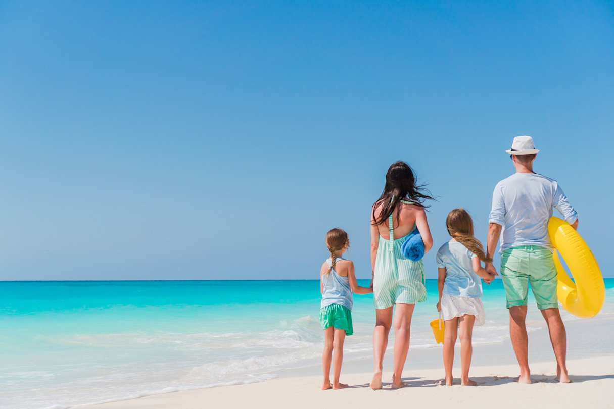 A young family standing on a sandy beach looking out to the clear blue sea on a hot day in summer