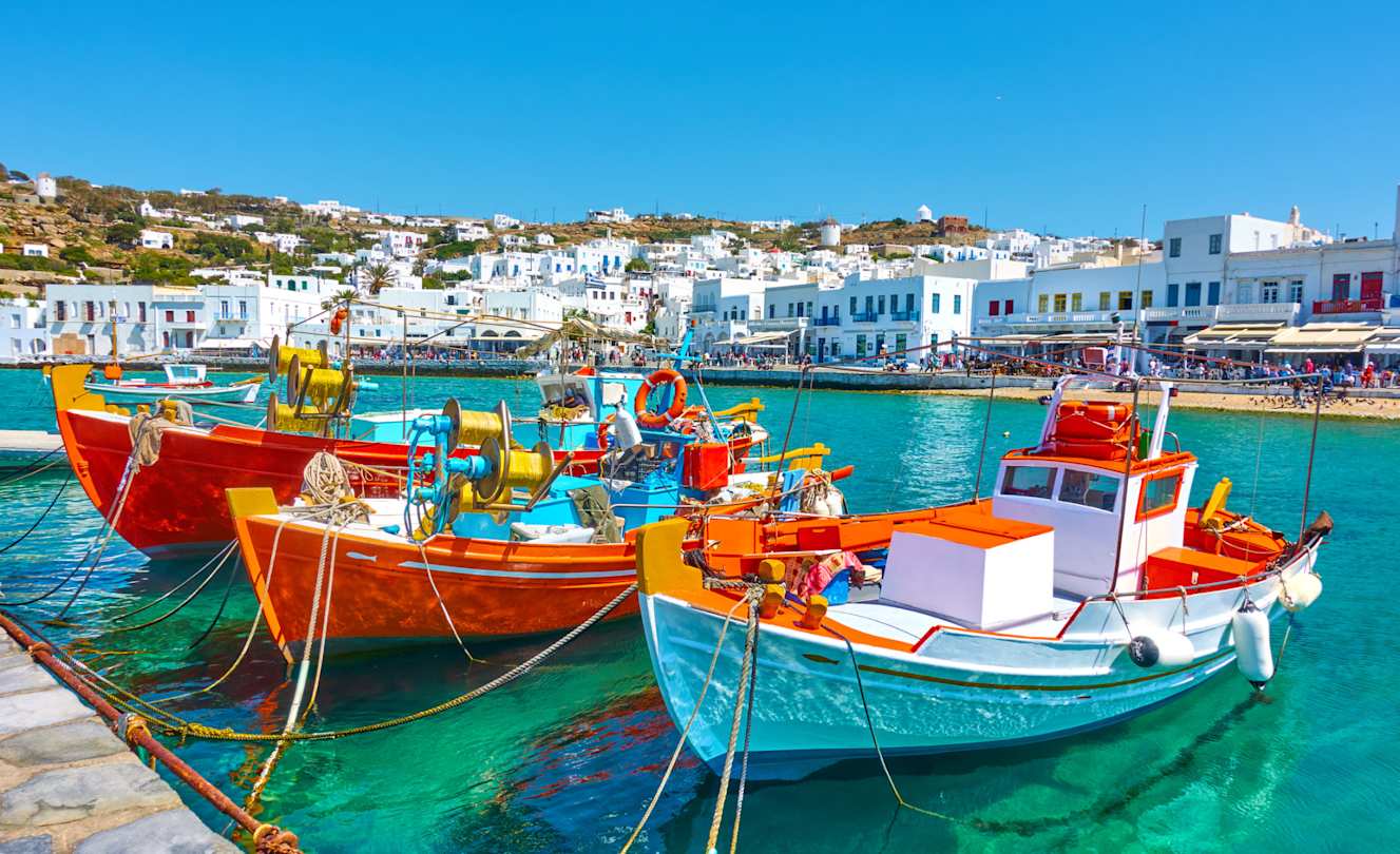 Three colourful boats docked on clear blue water in front of shops in Mykonos Town, Greece