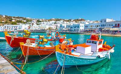 Three colourful boats docked on clear blue water in front of shops in Mykonos Town, Greece