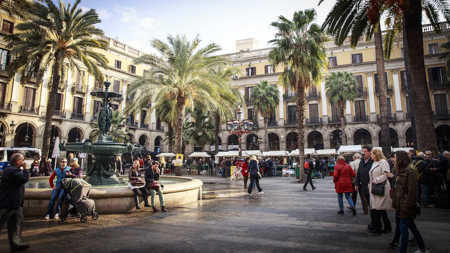 People among palm trees, buildings and a water fountain in a typical square in Barcelona, Spain 
