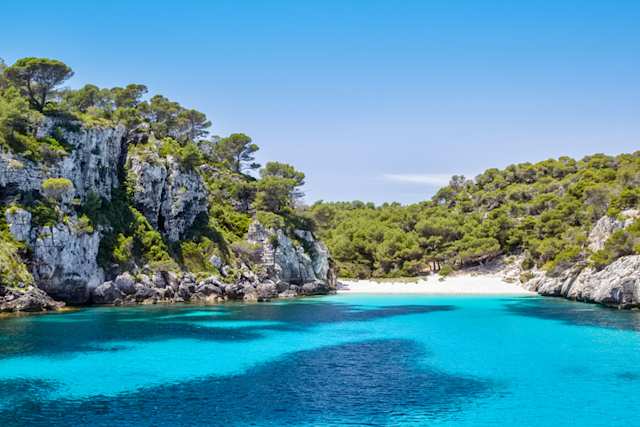 A view of bright blue water surrounded by grey cliffs and green trees at Cala Macarelleta Beach, Menorca, Spain