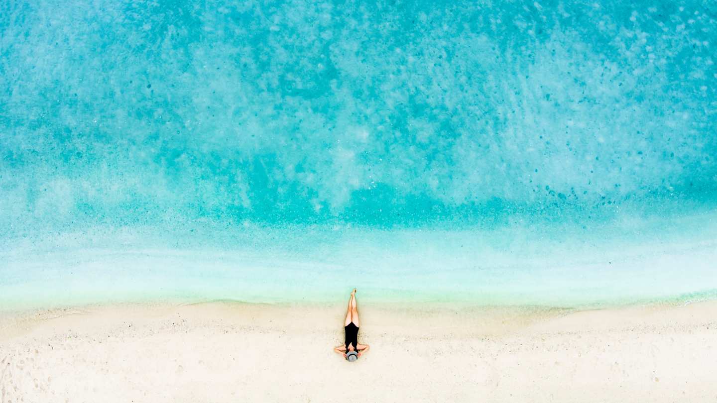 A bird's eye view of a woman sunbathing on a quiet, peaceful beach with clear blue water and white sand