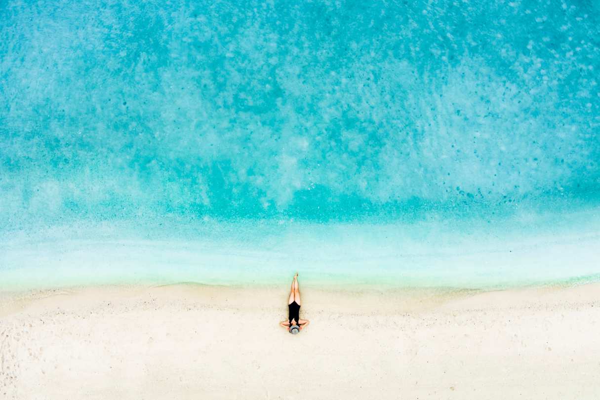 A bird's eye view of a woman sunbathing on a quiet, peaceful beach with clear blue water and white sand