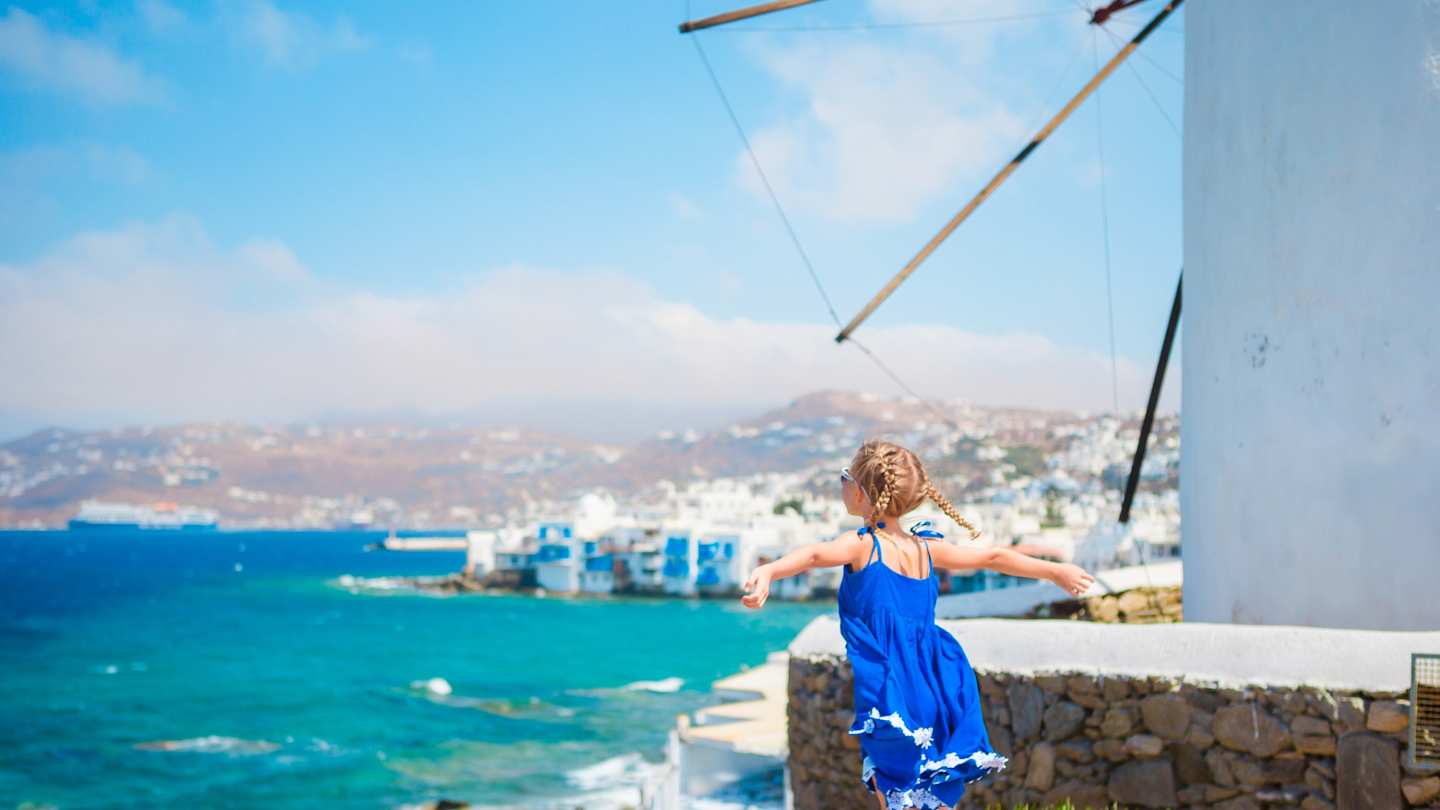 A young girl in a joyful pose next to a windmill looking out to sea over Mykonos, Greece