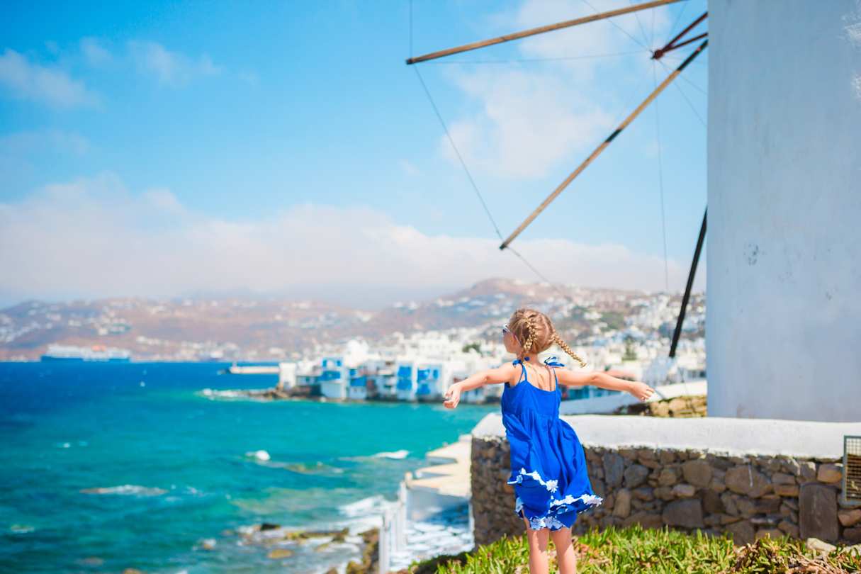 A young girl in a joyful pose next to a windmill looking out to sea over Mykonos, Greece