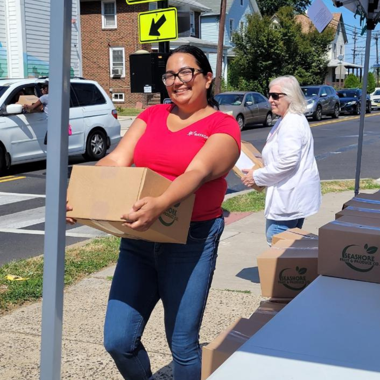 Woman in red BAYADA t-shirt holding a cardboard box of supplies, stands smiling and posed for the camera