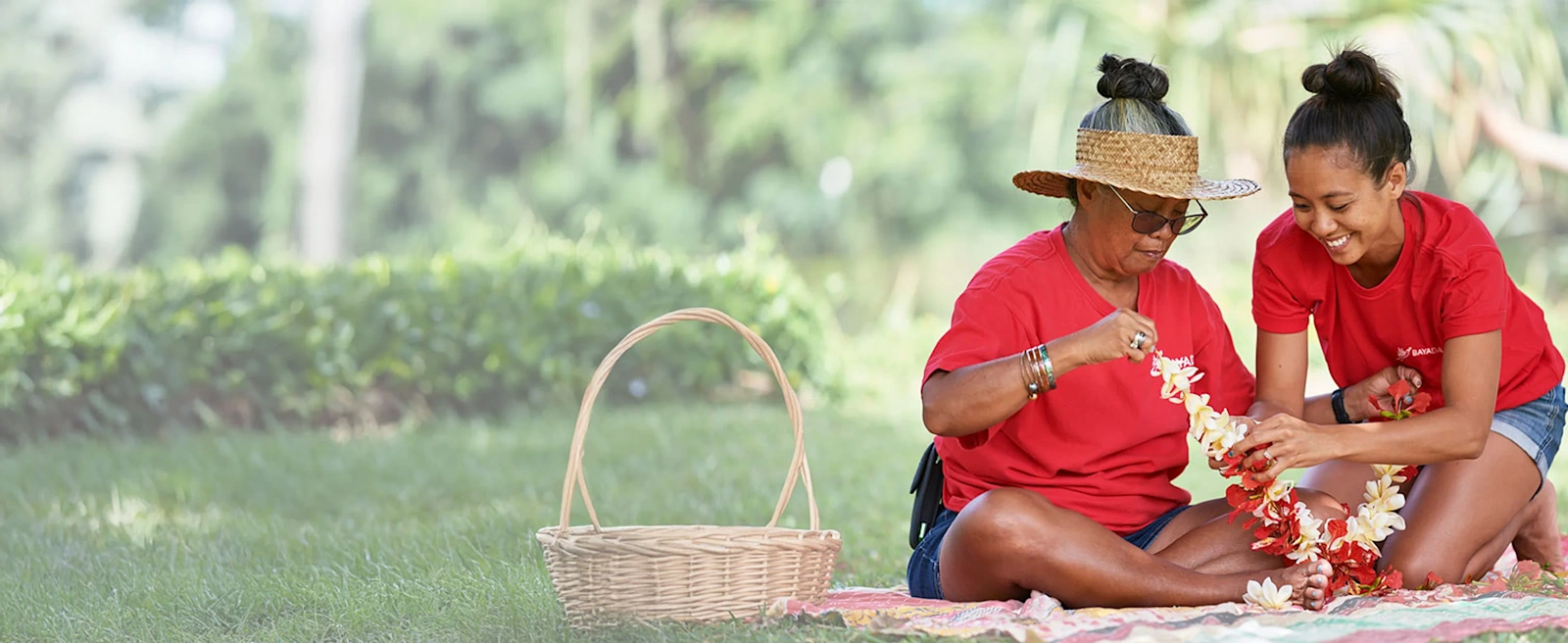 Two BAYADA Caregivers sit on a blanket outdoors, crafting flower leis.