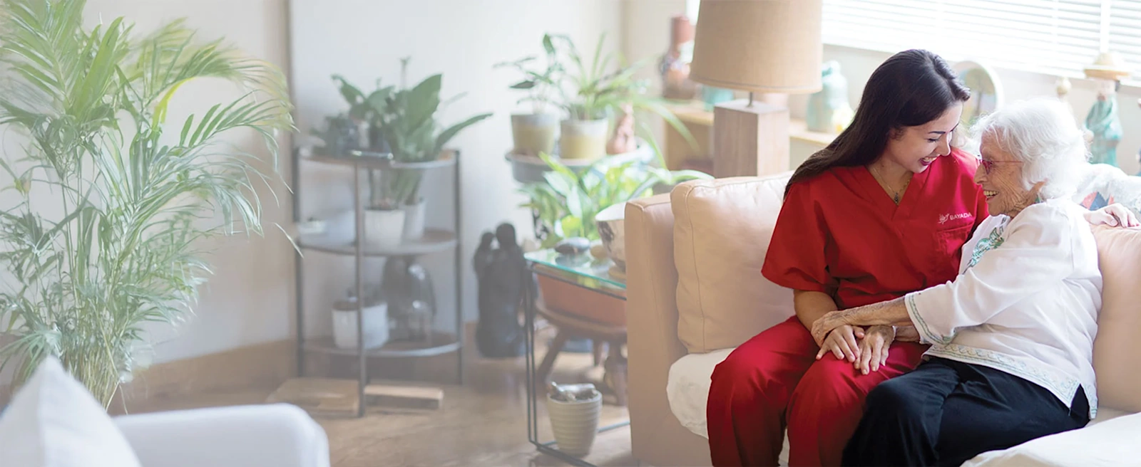 A BAYADA caregiver in red scrubs and an elderly woman sit on a cozy sofa, smiling warmly at each other in a bright room filled with houseplants.