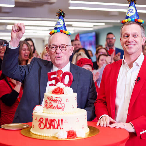 Two men celebrate a 50th anniversary with a cake at an office event.