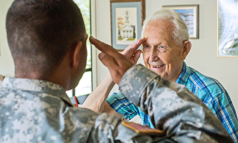 Soldier in uniform salutes an elderly man in a plaid shirt indoors. The man smiles warmly, evoking a sense of respect and camaraderie. BAYADA's Veteran Services Salute to Service.