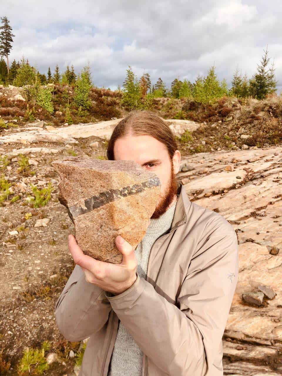 A picture of Jeroen Kortekaas holding a granite rock with a line of quartz veined through it, juxtaposed in front of his face, his right eye showing.