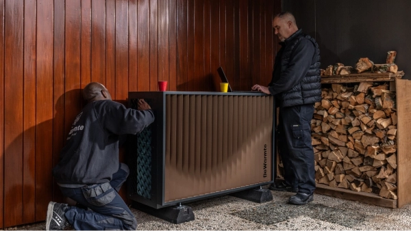DeWarmte Pomp MP All electric ready warmtepomp buitenunit wordt door twee installateurs tegen de gevel naast het houthok geplaatst