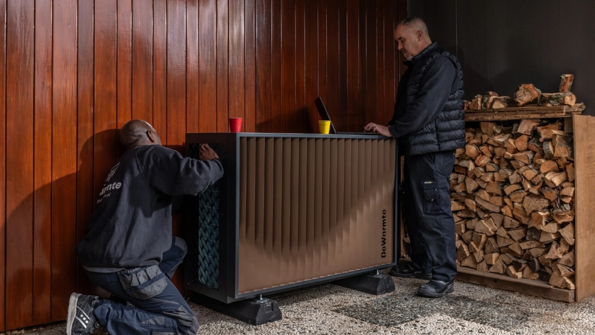 DeWarmte Pomp MP All electric ready warmtepomp buitenunit wordt door twee installateurs tegen de gevel naast het houthok geplaatst