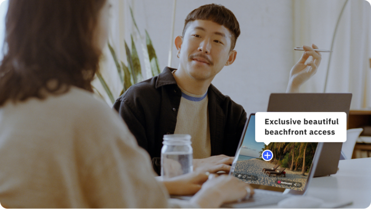 A man and a woman talking to each other at a table. Each has a laptop. The woman's laptop shows a tropical Matterport model with a note that says "Exclusive beautiful beachfront access"
