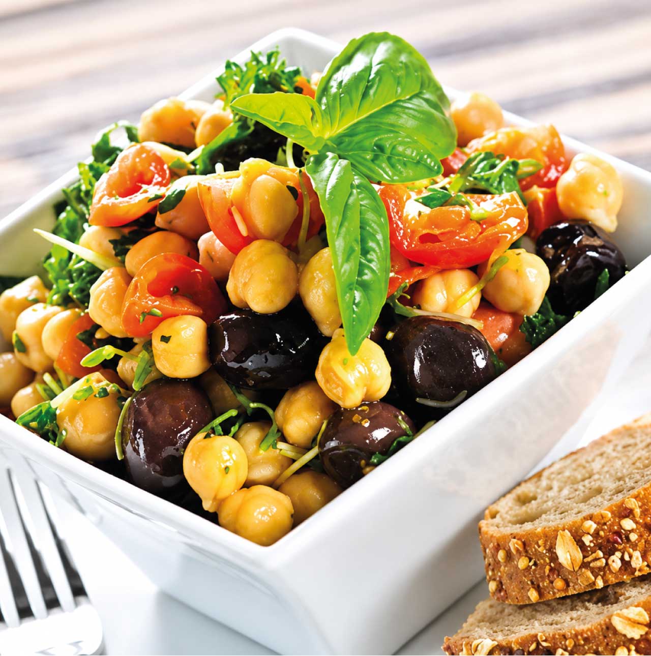 Plato de ensalada de verano con garbanzos cocidos, tomates cherry cortados, aceitunas negras y hojas de albahaca fresca en una mesa de picnic, receta infantil de Alcampo