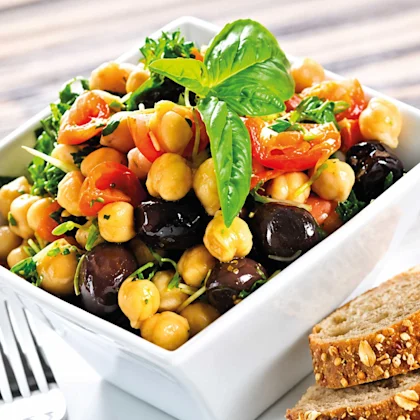 Plato de ensalada de verano con garbanzos cocidos, tomates cherry cortados, aceitunas negras y hojas de albahaca fresca en una mesa de picnic, receta infantil de Alcampo