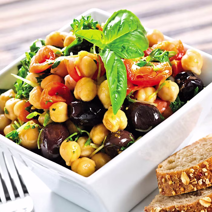 Plato de ensalada de verano con garbanzos cocidos, tomates cherry cortados, aceitunas negras y hojas de albahaca fresca en una mesa de picnic, receta infantil de Alcampo