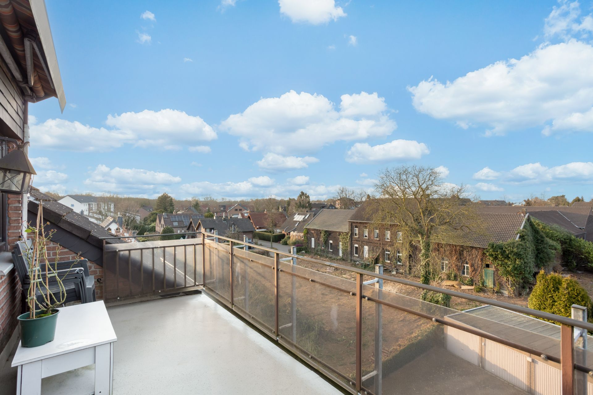 Balkon mit Glasgeländer und Blick auf Nachbarhäuser und blauen Himmel mit Wolken.