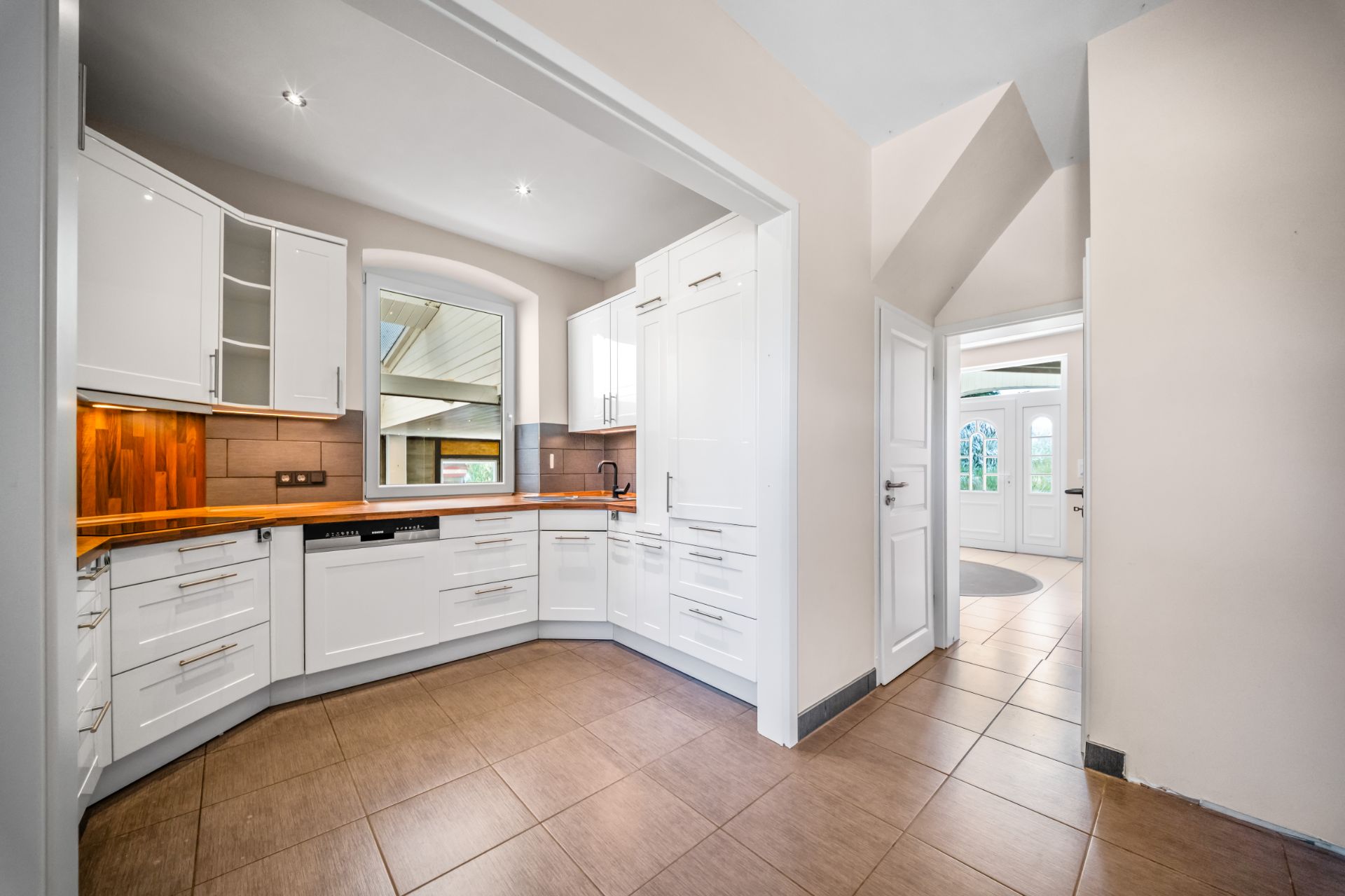 Moderne, helle Einbauküche mit weißen Schränken und Holzarbeitsplatte, Fenster und Flur im Hintergrund.