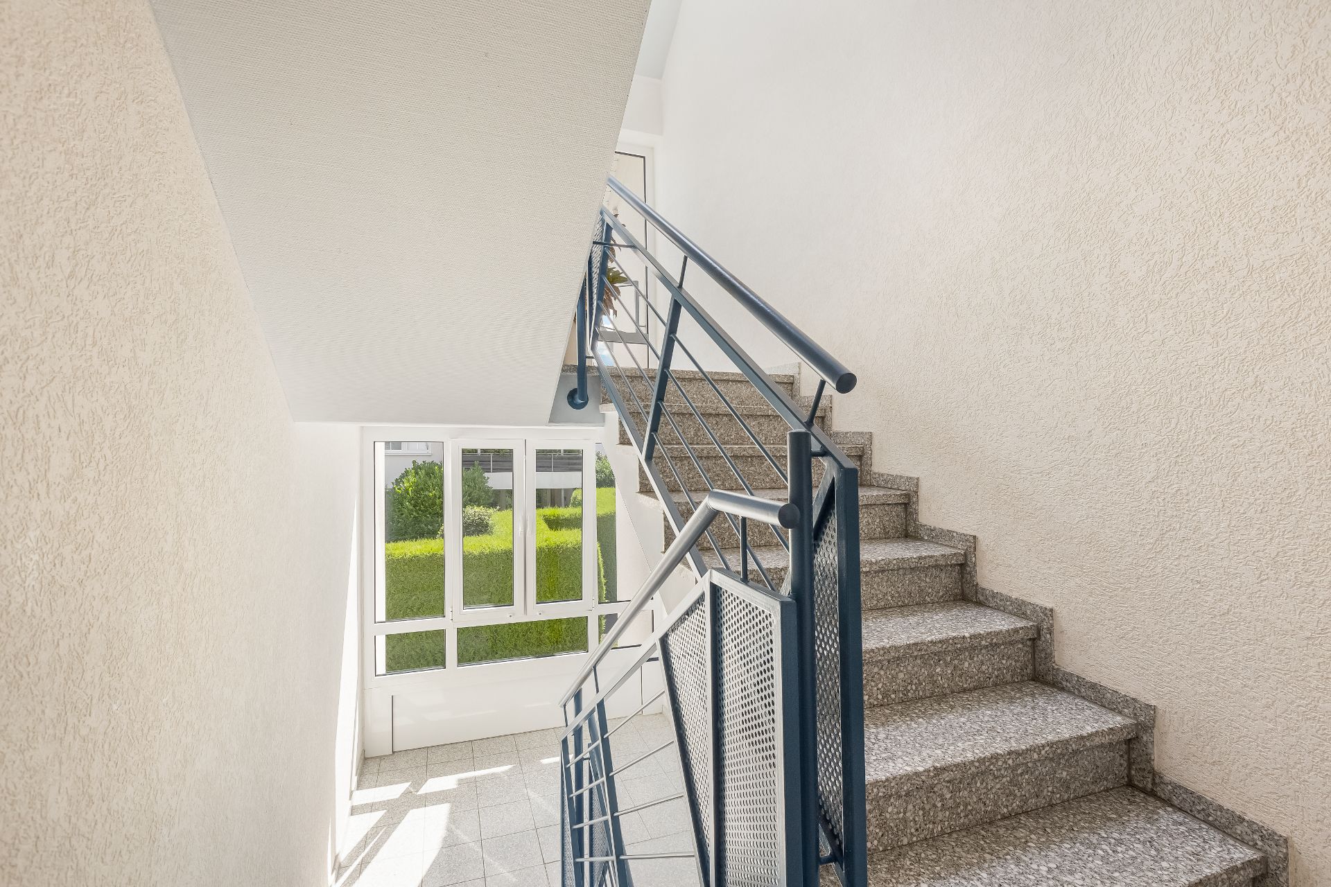 Helles Treppenhaus mit grauen Stufen und Metallgeländer, Fenster mit Blick ins Grüne.