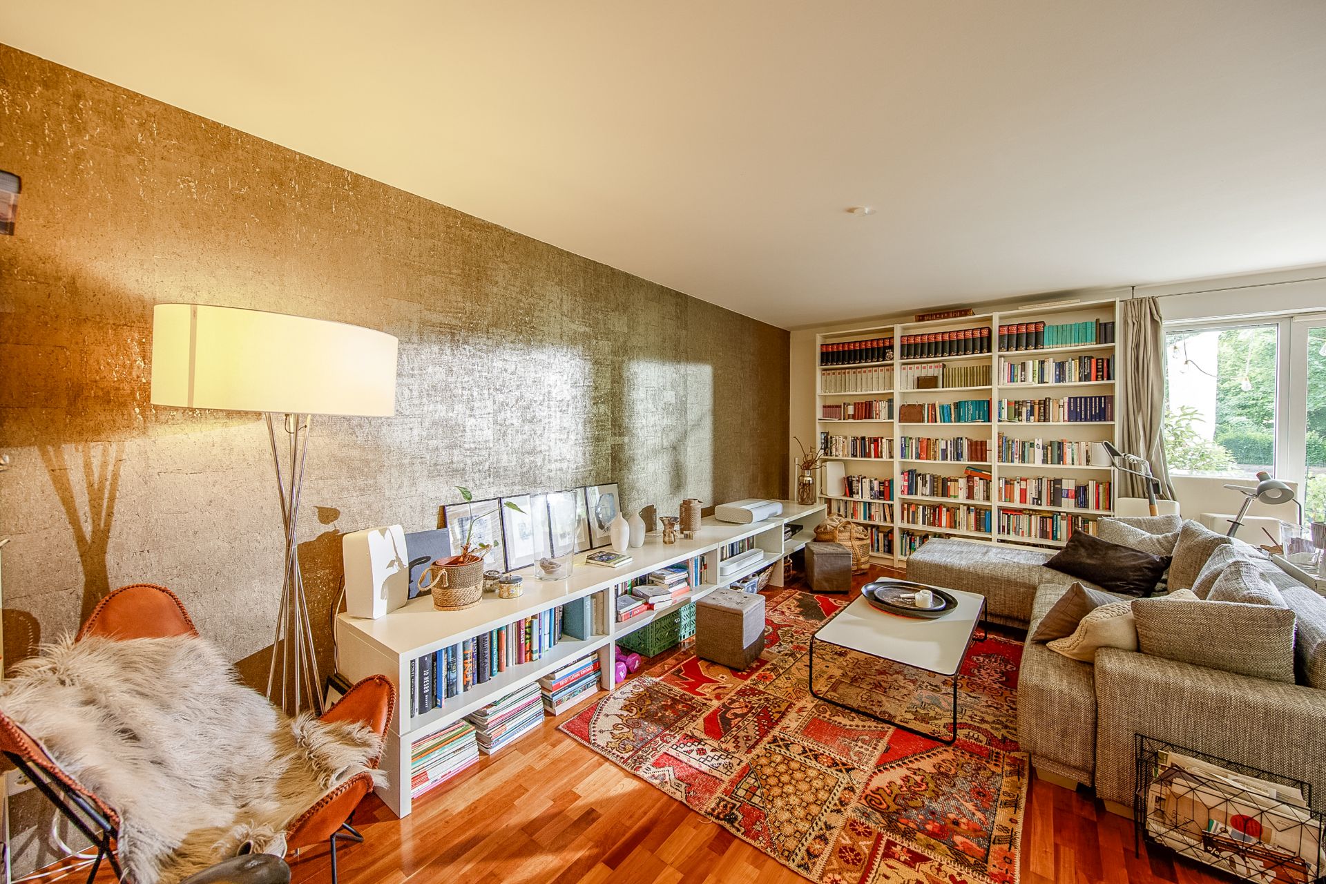 Wohnzimmer mit Sofa, Bücherregalen, Stehlampe und buntem Teppich vor großem Fenster.