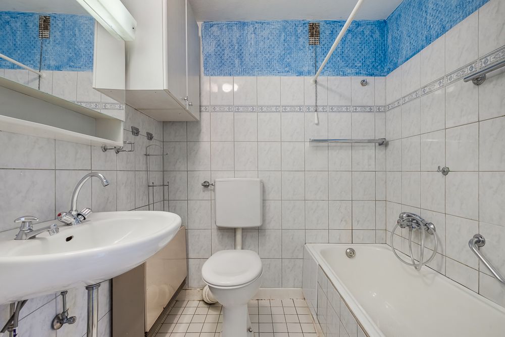 Kleines Badezimmer mit Waschbecken, Toilette und Badewanne, weiße Fliesen und blaue Wand.
