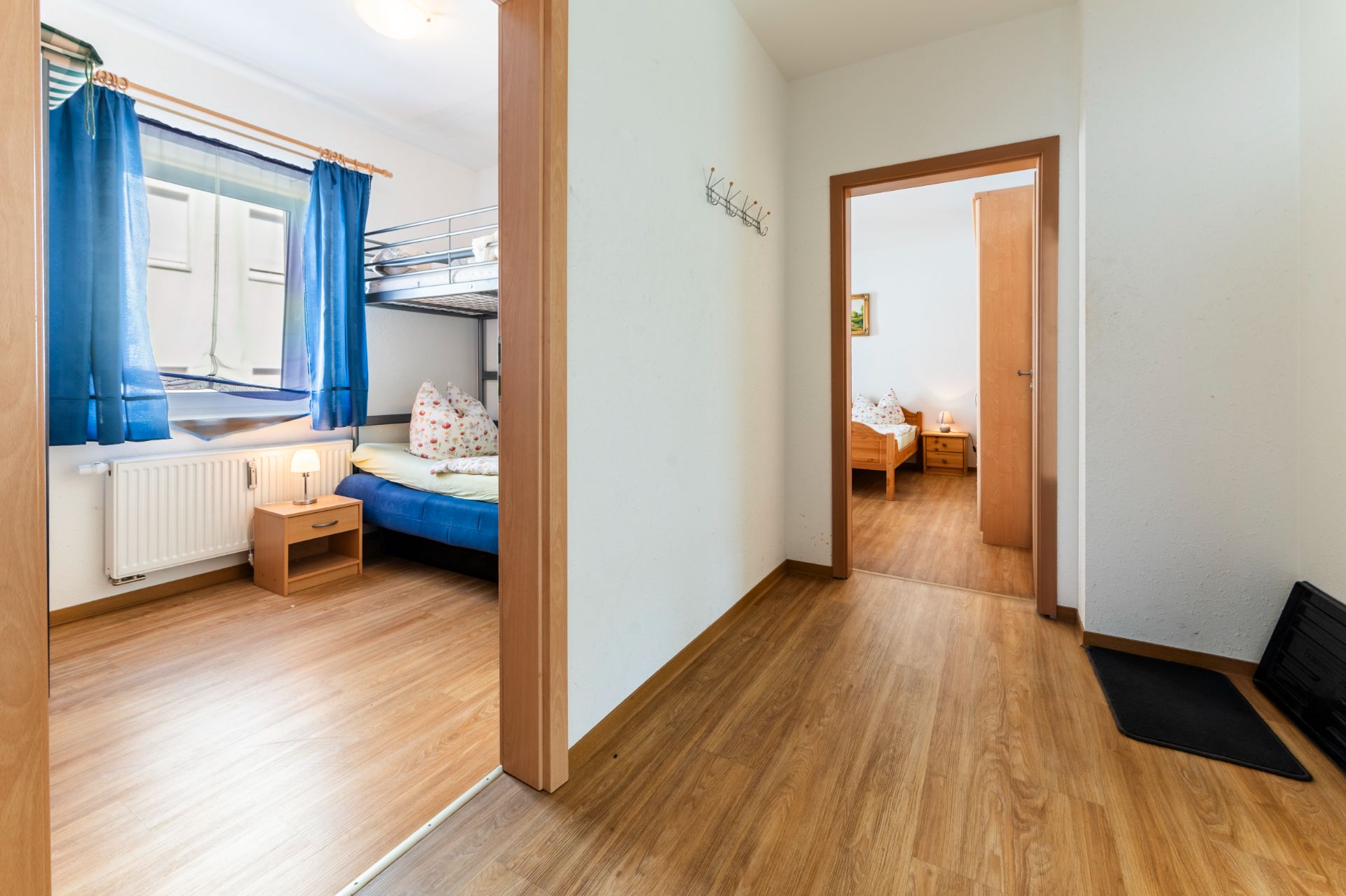 Flur mit Holzboden, Zugang zu zwei Schlafzimmern mit Betten und Nachttischen, blaue Vorhänge am Fenster.