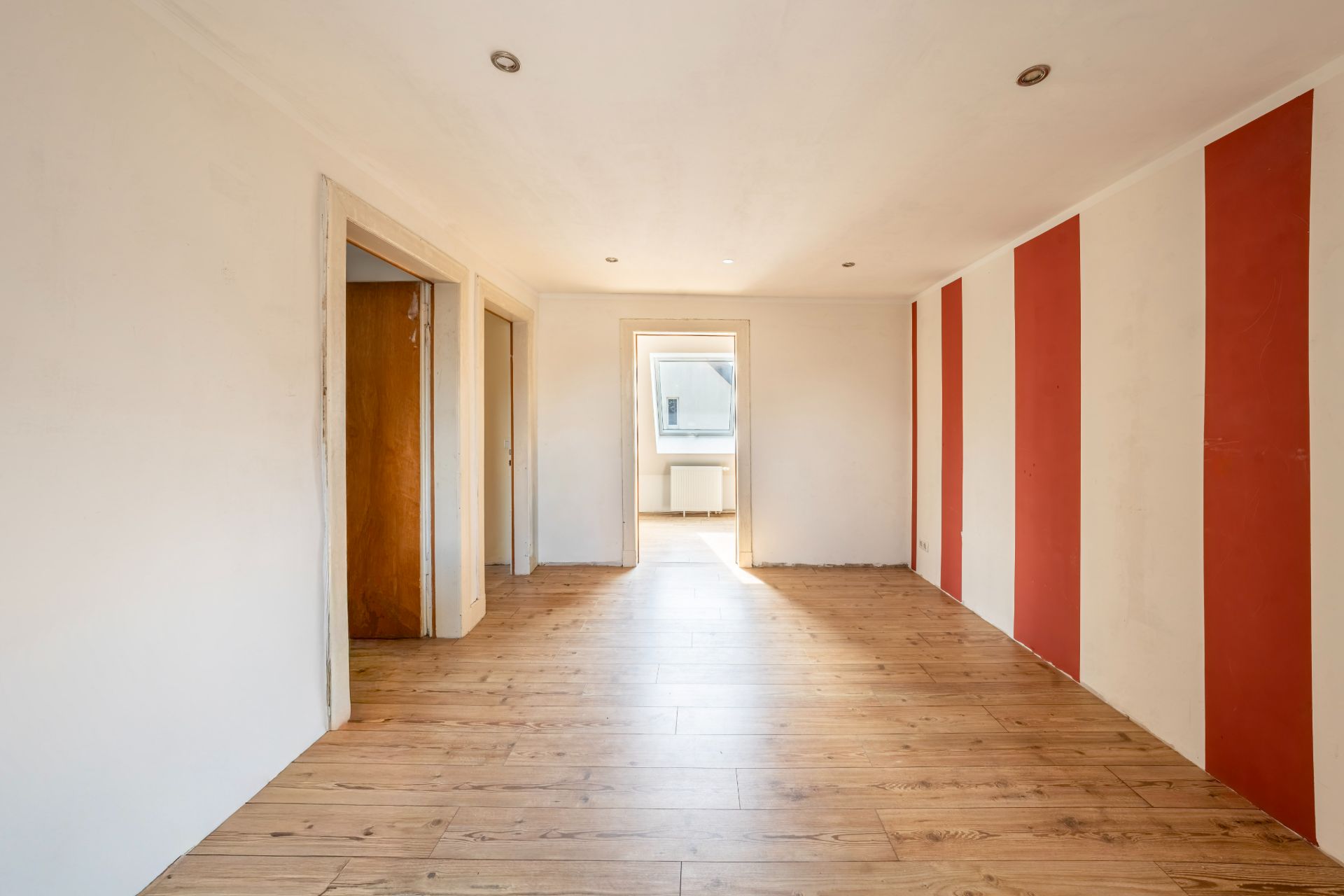 Leerer Raum mit Holzboden und rot-weiß gestreifter Wand, Türen führen zu weiteren Zimmern.