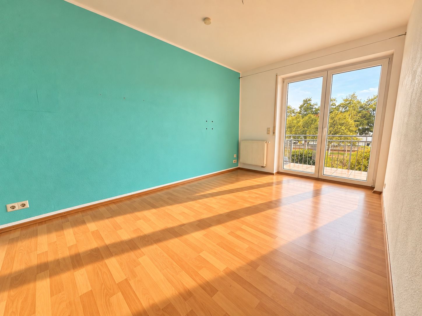 Helles, leeres Zimmer mit Holzfußboden, türkisfarbener Wand und großer Fensterfront mit Blick ins Grüne.