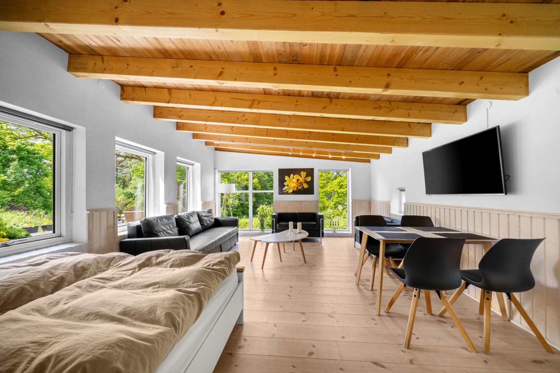 Helles Wohnzimmer mit Holzbalkendecke, Sofa, Esstisch, TV und Bett, große Fenster mit Blick ins Grüne.