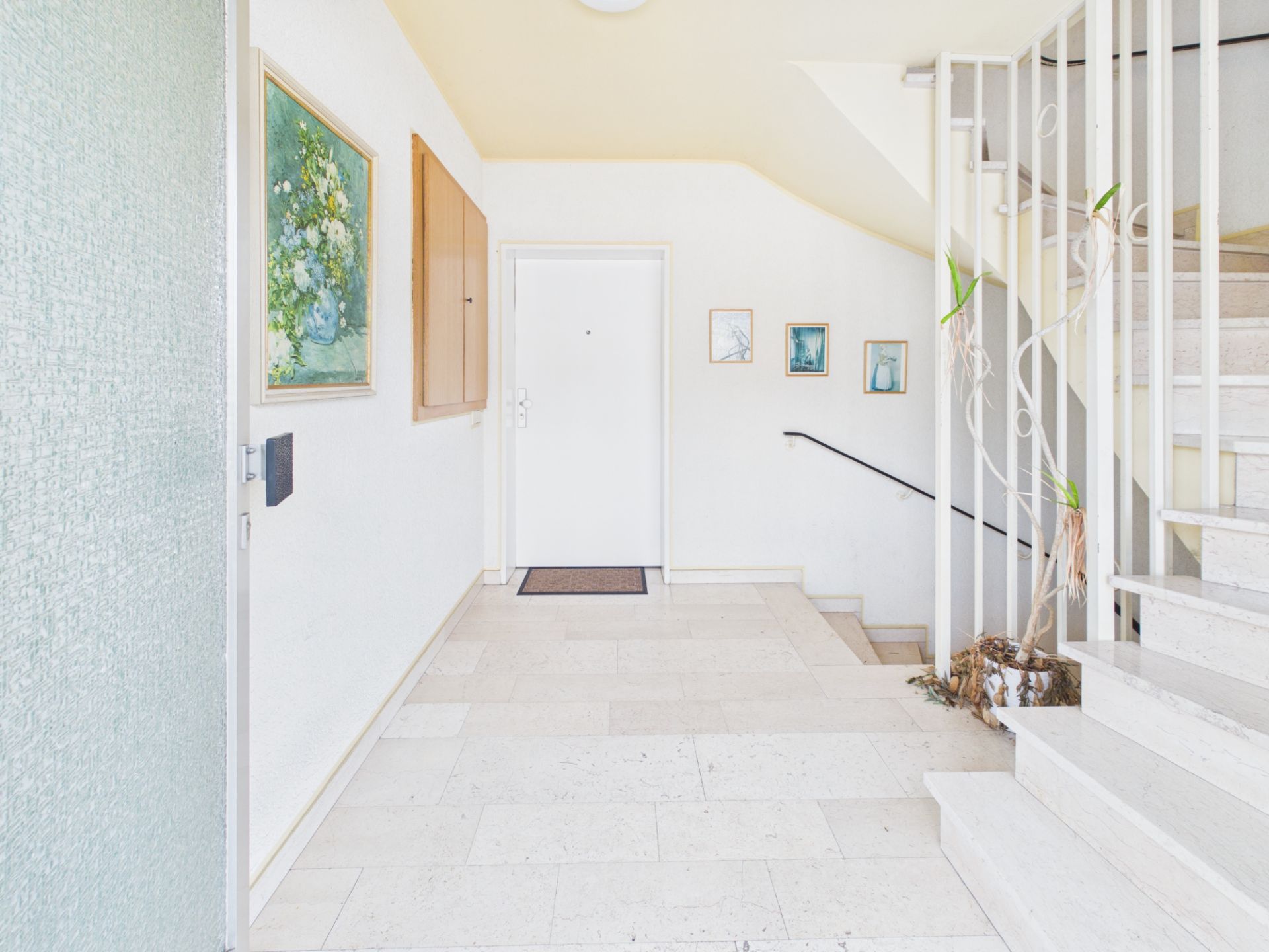 Heller Treppenhausflur mit weißen Wänden, Gemälden, einer Tür und einer Pflanze neben der Treppe.